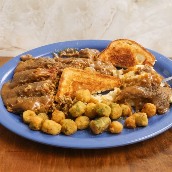 Blue plate piled with gravy-covered meat, mashed potatoes, stuffing, toasted bread, and fried vegetables on a wooden table.
