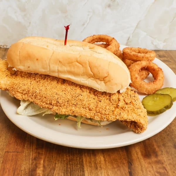 Fried fish sandwich on a hoagie roll with lettuce, onion rings, and pickle slices on a plate.